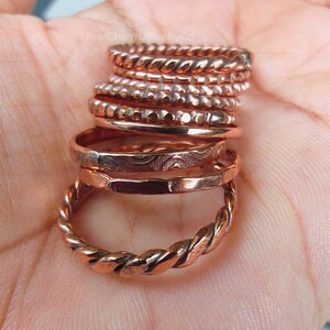 Puede incluir: Una colecci&oacute;n de anillos de color cobre con varios dise&ntilde;os, incluyendo trenzados, con cuentas y grabados. Los anillos est&aacute;n apilados, mostrando diferentes texturas y estilos. La imagen est&aacute; bien iluminada, resaltando el brillo met&aacute;lico de las joyas.
