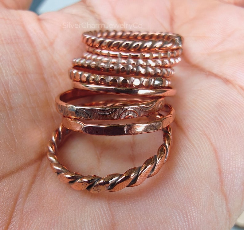 Puede incluir: Una colecci&oacute;n de anillos de color cobre con varios dise&ntilde;os, incluyendo trenzados, con cuentas y grabados. Los anillos est&aacute;n apilados, mostrando diferentes texturas y estilos. La imagen est&aacute; bien iluminada, resaltando el brillo met&aacute;lico de las joyas.