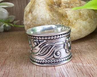 Könnte beinhalten: Ein silberner Ring mit floralen und Blattmustern. Der breite Ring hat detaillierte Gravuren von Blumen, Blättern und Wirbelmustern. Der Ring ist vor einem natürlichen Hintergrund platziert.