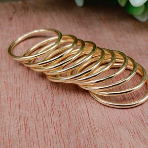 Pode incluir: Uma coleção de anéis finos dourados dispostos sobre uma superfície de madeira. Os anéis são empilhados, mostrando seu design simples e elegante. Os anéis parecem ser de metal e têm um acabamento liso e polido. O fundo é uma madeira quente, marrom-avermelhada.