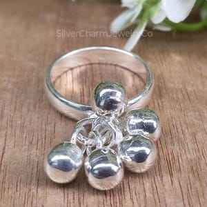 Puede incluir: Un anillo de plata con un grupo de pequeños dijes redondos colgantes. El anillo y los dijes están muy pulidos, reflejando la luz. Los dijes están unidos al anillo con delicados eslabones. El anillo está sobre una superficie de madera.