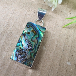 Puede incluir: Colgante rectangular con incrustación de concha de abulón iridiscente en tonos azules, verdes y morados. La concha está engastada en un marco de metal plateado con una anilla a juego. Una pieza de joyería única.