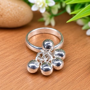 May include: A polished silver ring featuring a cluster of five small, round, metallic ball charms suspended from the band by individual jump rings. The band is smooth and circular, and the charms are reflective, creating a shiny, metallic appearance.