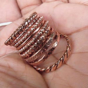Puede incluir: Un conjunto de anillos de color cobre con diversos dise&ntilde;os, incluyendo trenzados, martillados y texturizados. Los anillos est&aacute;n apilados, mostrando una variedad de estilos y anchos. La imagen es un primer plano, destacando los detalles de la joyer&iacute;a.