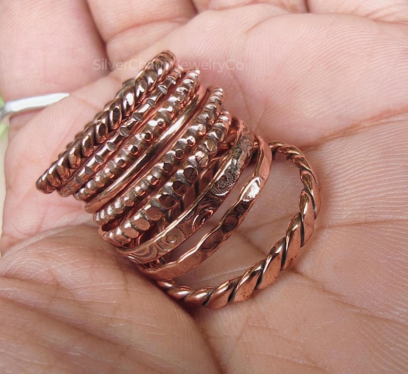 Puede incluir: Un conjunto de anillos de color cobre con diversos dise&ntilde;os, incluyendo trenzados, martillados y texturizados. Los anillos est&aacute;n apilados, mostrando una variedad de estilos y anchos. La imagen es un primer plano, destacando los detalles de la joyer&iacute;a.