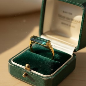 Könnte beinhalten: Ein goldener Ring mit einem rechteckigen, teal-farbenen Edelstein, präsentiert in einer offenen, dunkelgrünen, samtgefütterten Schmuckschatulle. Der Ring hat ein goldenes Band. Die Schmuckschatulle ist im Vintage-Stil mit einem goldenen Verschluss. Der Text "The Aurielle Atelier" ist sichtbar.