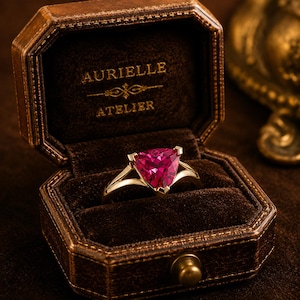 May include: A gold ring with a triangular, vibrant pink gemstone, displayed in a brown leather jewelry box. The box is lined with dark brown velvet and has the text "AURIELLE ATELIER" embossed on the inside.