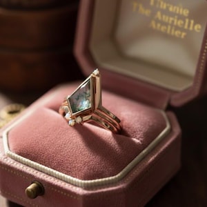 Puede incluir: Un anillo de oro rosa con un diseño geométrico, con una piedra verde angular grande con detalles en blanco. El anillo se muestra en un joyero de terciopelo rosa. La caja tiene el texto "The Aurielle Atelier".