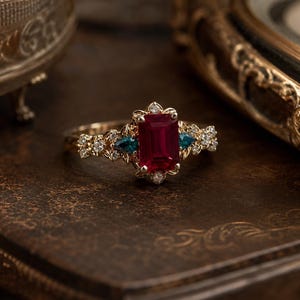 May include: A gold ring featuring a central emerald-cut red gemstone flanked by two small pear-cut teal stones. The band is decorated with intricate leaf-like metalwork and small round white accent stones surrounding the center setting.