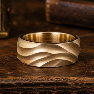 May include: A gold-colored ring with a wide band featuring a textured, undulating surface that mimics the appearance of rolling sand dunes. The metal has a matte finish, and the interior of the band is smooth and polished, showing a reflective surface.