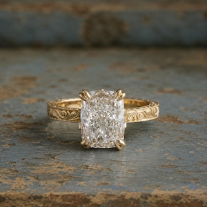 May include: A gold ring featuring a large, rectangular cushion-cut clear gemstone held by four prongs. The band is crafted from yellow gold and displays an intricate, engraved floral scroll pattern along its entire outer surface.