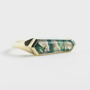 Puede incluir: Anillo de oro con una gema rectangular facetada con inclusiones verdes y beige. El anillo presenta un diseño moderno y sencillo con una banda y engaste de oro pulido. Los patrones de la gema evocan un paisaje en miniatura.