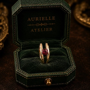 May include: A gold ring with a vibrant red gemstone, presented in a dark green velvet jewelry box. The box is embossed with the words "AURIELLE ATELIER". The ring's design features a split shank, showcasing the gemstone.