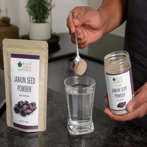 Puede incluir: Una bolsa de papel marrón y un frasco de plástico transparente de polvo de semillas de jamun Bliss of Earth, con un logo de hoja verde y una imagen de jamun. Una cuchara de plata sostiene polvo marrón sobre un vaso de agua transparente.