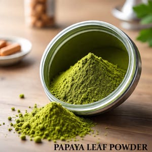 May include: A close-up of a silver tin filled with bright green papaya leaf powder, with a small pile of the powder spilled out onto a wooden surface. The text "PAPAYA LEAF POWDER" is printed at the bottom of the image.