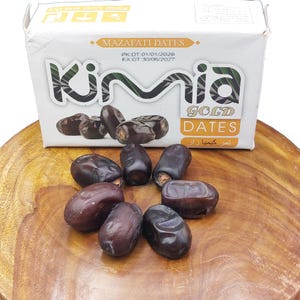 May include: A white box of Kimia Gold Dates with the text "MAZAFATI DATES" and "GOLD DATES" in gold lettering. Several dark brown dates are scattered in front of the box on a wooden surface. The box has a production and expiration date.