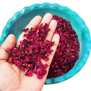 Pode incluir: Close-up de pétalas de rosa secas, vermelho escuro, seguradas em uma mão, com mais pétalas em uma tigela turquesa. As pétalas têm uma aparência texturizada e várias tonalidades de vermelho e amarelo.