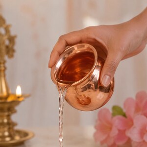 Puede incluir: Un pequeño recipiente redondo de color cobre se inclina para verter un chorro de líquido transparente. El recipiente tiene una abertura ancha y una superficie lisa y reflectante. Una lámpara encendida y flores rosas están en el fondo.