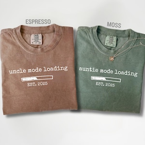 May include: Two folded t-shirts, one espresso brown and one moss green. The espresso shirt reads "uncle mode loading" with a progress bar and "EST. 2025". The green shirt says "auntie mode loading" with the same design. Both shirts have a crew neck.