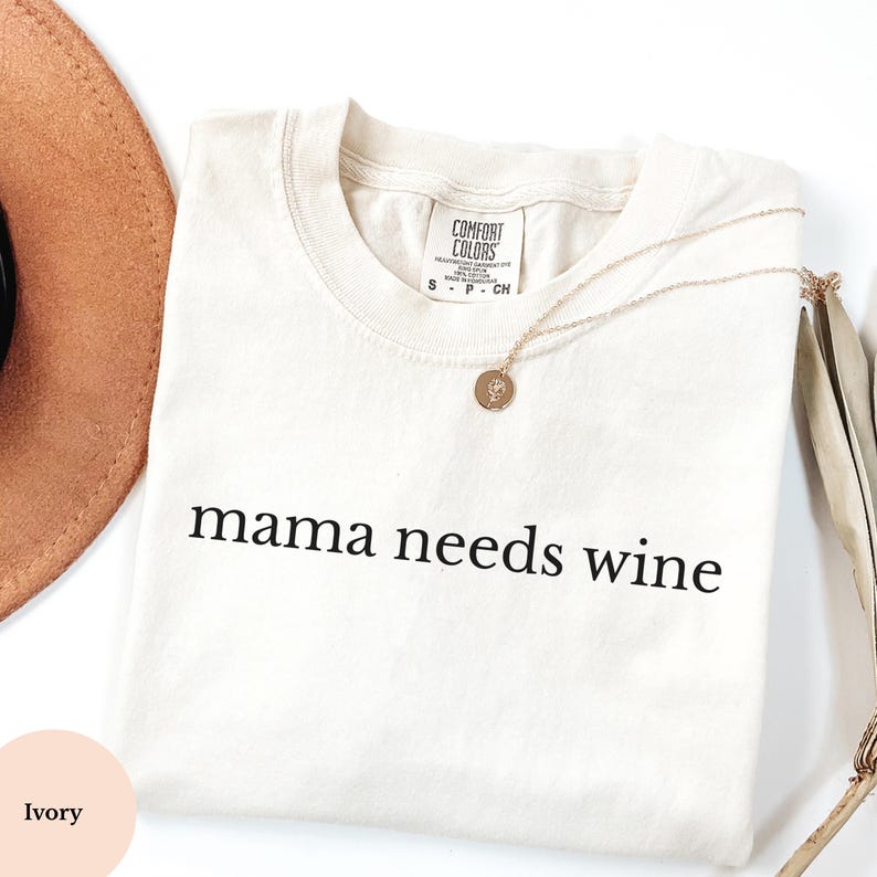 May include: Cream-coloured t-shirt with the words "mama needs wine" printed in black. A gold necklace with a pendant and a brown hat are also visible. The shirt is from Comfort Colors.