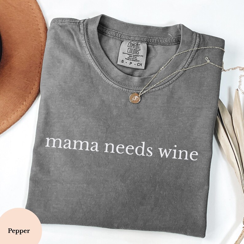 May include: A grey Comfort Colors t-shirt with the words "mama needs wine" printed in white. A silver necklace with a pendant lies on the shirt. A brown hat and decorative leaves are in the background.