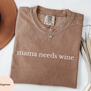 May include: Espresso-coloured t-shirt with the phrase "mama needs wine" printed in white. A silver necklace with a pendant rests on the shirt. The shirt is folded and has a Comfort Colors tag.