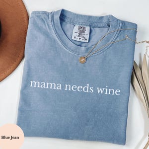 May include: A blue jean coloured t-shirt with the phrase "mama needs wine" printed in white. A gold necklace with a pendant rests on the shirt. The shirt is folded on a white surface.