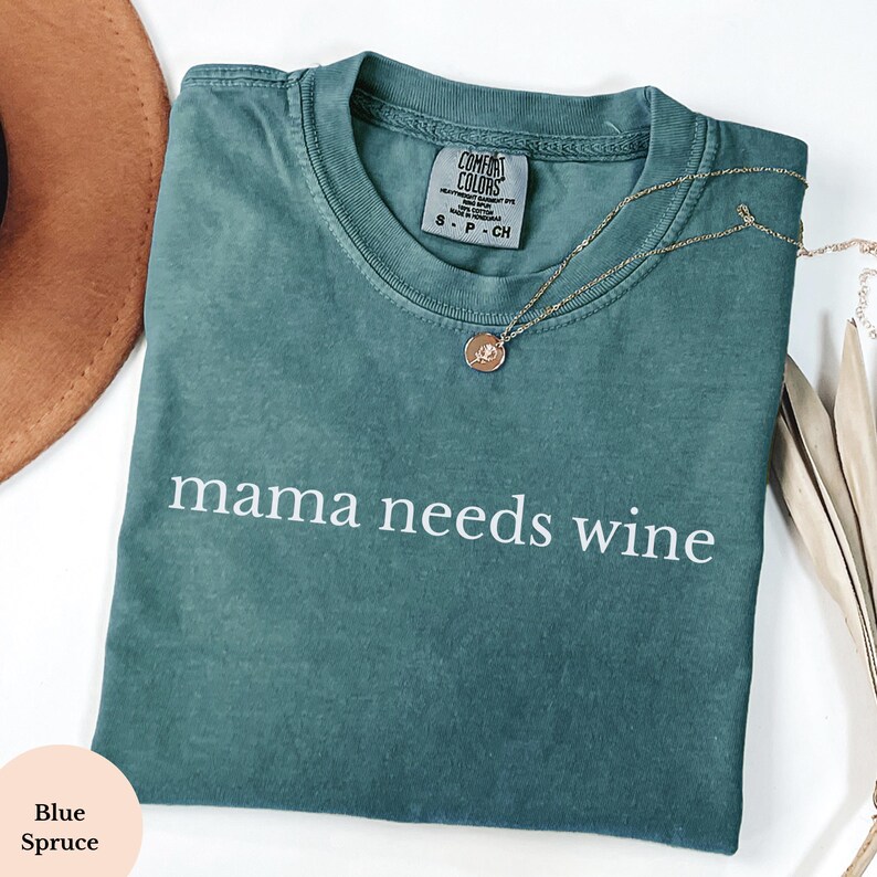 May include: A teal-coloured t-shirt with the words "mama needs wine" printed in white. The shirt has a crew neck and short sleeves. A gold necklace with a pendant is draped across the shirt. The shirt is folded on a white surface.