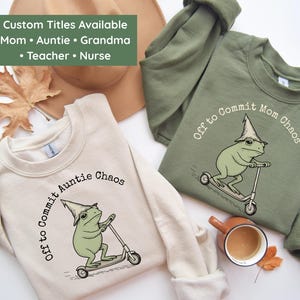 May include: Two sweatshirts, one cream and one olive green, with a frog graphic riding a scooter. The cream sweatshirt says "Off to Commit Auntie Chaos." The green sweatshirt says "Off to Commit Mom Chaos." Text at the top says "Custom Titles Available: Mom, Auntie, Grandma, Teacher, Nurse."