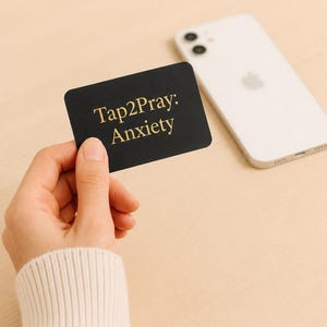 Può includere: Una carta rettangolare nera con angoli arrotondati che presenta il testo dorato Tap2Pray: Anxiety stampato sul davanti. La carta è tenuta in una mano, posizionata vicino a uno smartphone bianco con un sistema a doppia fotocamera sul retro.