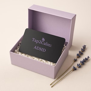 May include: A black card with the text "Tap2Calm: ADHD" inside a lavender gift box with white paper filler. Two sprigs of lavender are placed next to the box. The box is open, revealing the card.