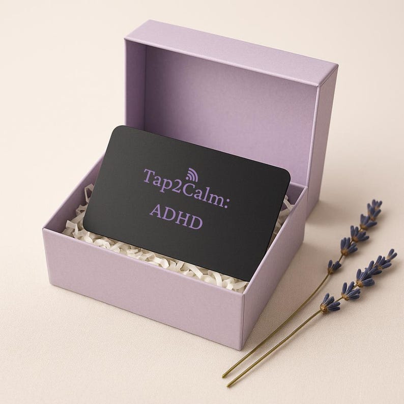 May include: A black card with the text "Tap2Calm: ADHD" inside a lavender gift box with white paper filler. Two sprigs of lavender are placed next to the box. The box is open, revealing the card.