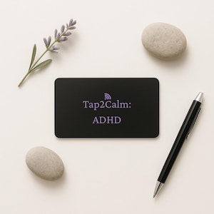 May include: A black rectangular card with the text "Tap2Calm: ADHD" in purple, a black pen, two smooth gray stones, and a sprig of lavender on a white surface.