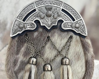 Può includere: Un tradizionale sporran scozzese con un cantle in metallo argentato con disegni di cardi e nodi celtici. Lo sporran ha un corpo in pelliccia con tre nappe pendenti e dettagli a catena. La tavolozza dei colori comprende argento, grigio e bianco.