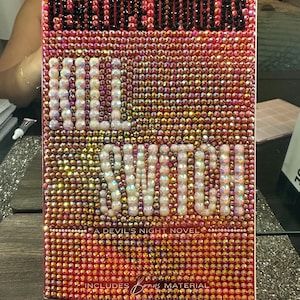 Pu&ograve; includere: Un libro intitolato "Kill Switch" di Penelope Douglas, ricoperto di strass rosa e oro scintillanti. Il titolo &egrave; in grandi perle bianche, simili a perle. La copertina include anche il testo "A Devil's Night Novel" e "Includes Bonus Material."