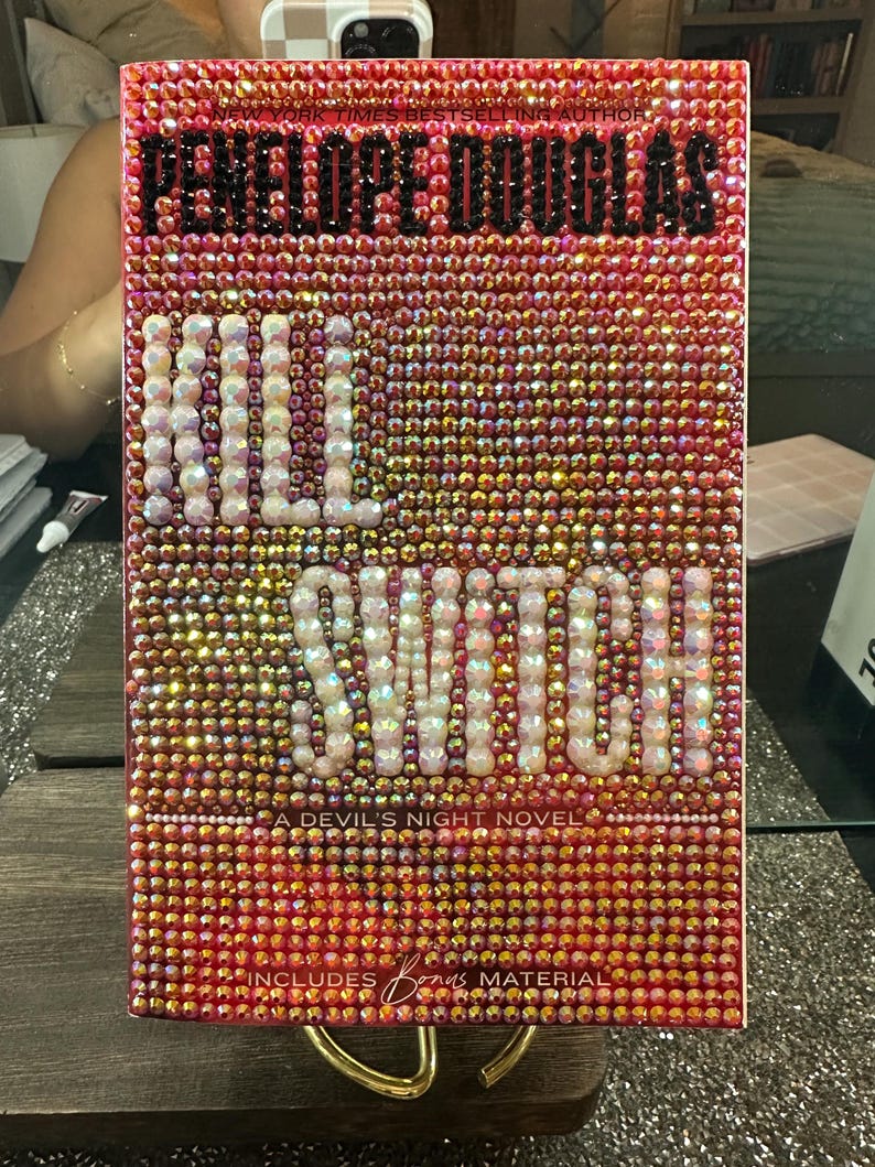 Pu&ograve; includere: Un libro intitolato "Kill Switch" di Penelope Douglas, ricoperto di strass rosa e oro scintillanti. Il titolo &egrave; in grandi perle bianche, simili a perle. La copertina include anche il testo "A Devil's Night Novel" e "Includes Bonus Material."