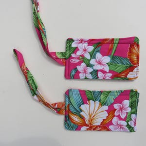 Peut inclure: Deux &eacute;tiquettes de bagages avec un motif floral vibrant. Les &eacute;tiquettes pr&eacute;sentent un fond rose vif avec des fleurs blanches et roses, des feuilles vertes et des accents orange. Chaque &eacute;tiquette a une sangle assortie pour l'attache.