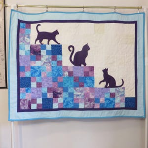 May include: A handmade quilt featuring three dark purple cat silhouettes on a white background. The quilt has a blue and purple patchwork design at the bottom, with a light blue border. The quilt is hanging on a white wall.
