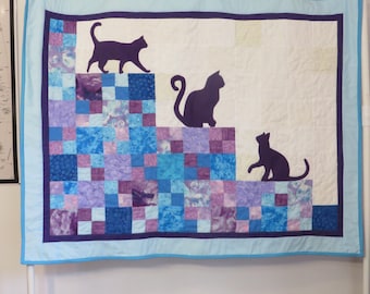 May include: A handmade quilt featuring three dark purple cat silhouettes on a white background. The quilt has a blue and purple patchwork design at the bottom, with a light blue border. The quilt is hanging on a white wall.