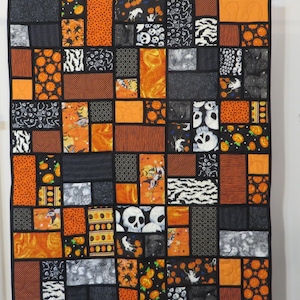 May include: A Halloween-themed quilt with a patchwork design. The quilt features various fabrics in black, orange, and white, with patterns including pumpkins, skulls, bats, and stripes. The quilt is rectangular and has a black border.