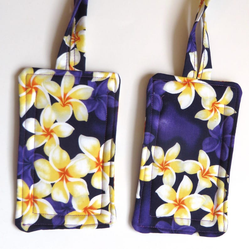 Peut inclure: Deux &eacute;tiquettes &agrave; bagages rectangulaires avec un motif floral. Les &eacute;tiquettes pr&eacute;sentent un fond bleu fonc&eacute; avec des fleurs de plumeria jaunes et blanches et des accents violets. Chaque &eacute;tiquette a une boucle en tissu assortie pour l'attache.