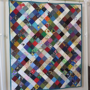 May include: A handmade quilt with a patchwork design. The quilt features a variety of colorful fabric squares arranged in a diagonal pattern. The squares include floral, geometric, and solid patterns in shades of blue, red, green, and brown. The quilt is trimmed with a teal border.