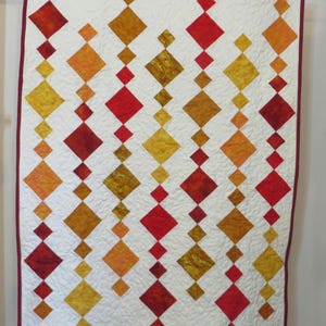 May include: A white quilt with a repeating pattern of red, orange, and yellow diamond shapes. The quilt is edged with a burgundy border.