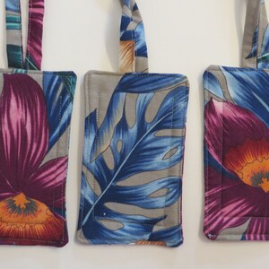 Peut inclure: Trois &eacute;tiquettes &agrave; bagages rectangulaires avec des motifs floraux. Les &eacute;tiquettes pr&eacute;sentent un fond gris avec des motifs vibrants de fleurs et de feuilles violettes, bleues et orange. Chaque &eacute;tiquette a une boucle en tissu assortie pour l'attache, et les bords sont cousus.