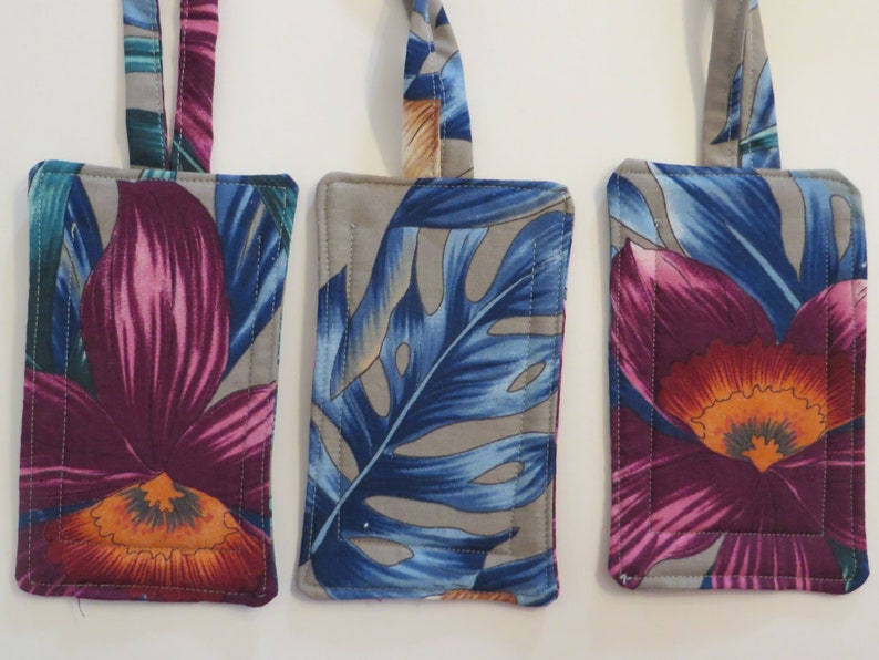Peut inclure: Trois &eacute;tiquettes &agrave; bagages rectangulaires avec des motifs floraux. Les &eacute;tiquettes pr&eacute;sentent un fond gris avec des motifs vibrants de fleurs et de feuilles violettes, bleues et orange. Chaque &eacute;tiquette a une boucle en tissu assortie pour l'attache, et les bords sont cousus.