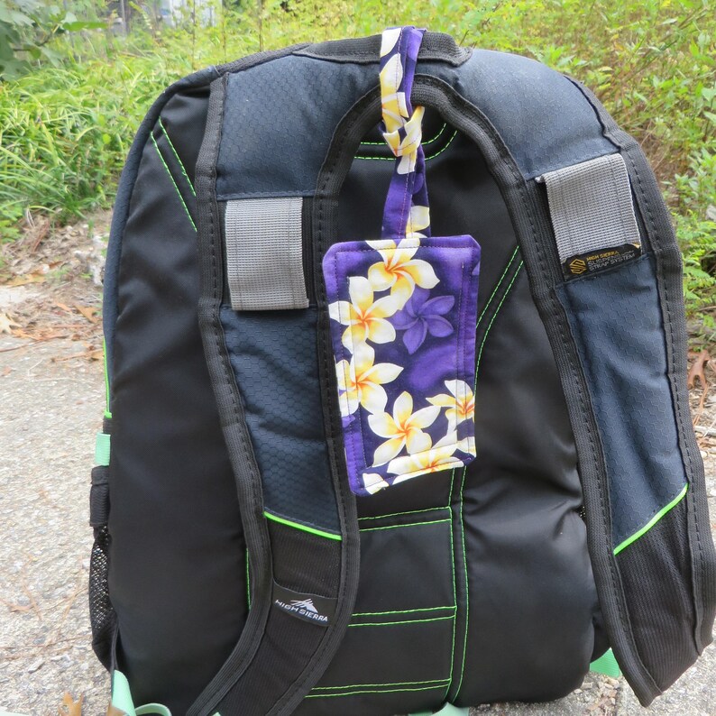 Peut inclure: Un sac &agrave; dos noir et bleu avec une &eacute;tiquette de bagage &agrave; motifs floraux. L'&eacute;tiquette pr&eacute;sente un fond violet avec des fleurs de plumeria blanches et jaunes. Le sac &agrave; dos a des accents verts et des bandes r&eacute;fl&eacute;chissantes.