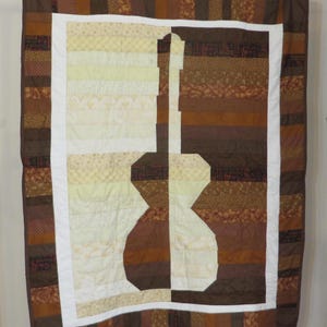 May include: A brown and cream patchwork quilt with a guitar silhouette in the center. The quilt is made of various fabrics with different patterns and textures.