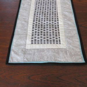 May include: A rectangular table runner with a musical theme. The center features a piano key pattern in black and white, framed by a cream border. The outer layer is gray with musical notes, and the edges are black and turquoise.