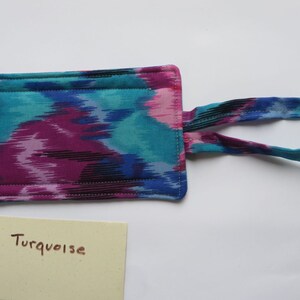 Peut inclure: Un article rectangulaire en tissu avec un motif ikat turquoise, magenta et noir vibrant. Il a une sangle assortie et est cousu avec du fil blanc. Une petite carte jaune clair avec le mot "Turquoise" est &eacute;galement pr&eacute;sente.