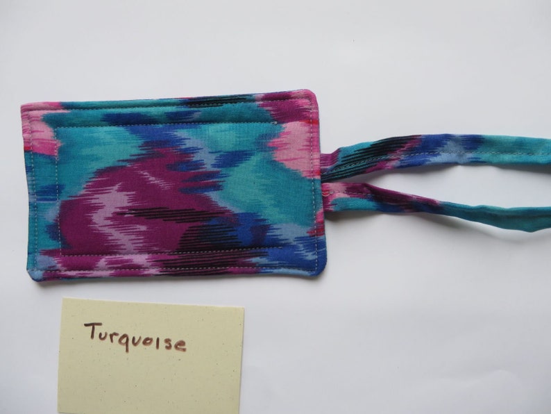 Peut inclure: Un article rectangulaire en tissu avec un motif ikat turquoise, magenta et noir vibrant. Il a une sangle assortie et est cousu avec du fil blanc. Une petite carte jaune clair avec le mot "Turquoise" est &eacute;galement pr&eacute;sente.